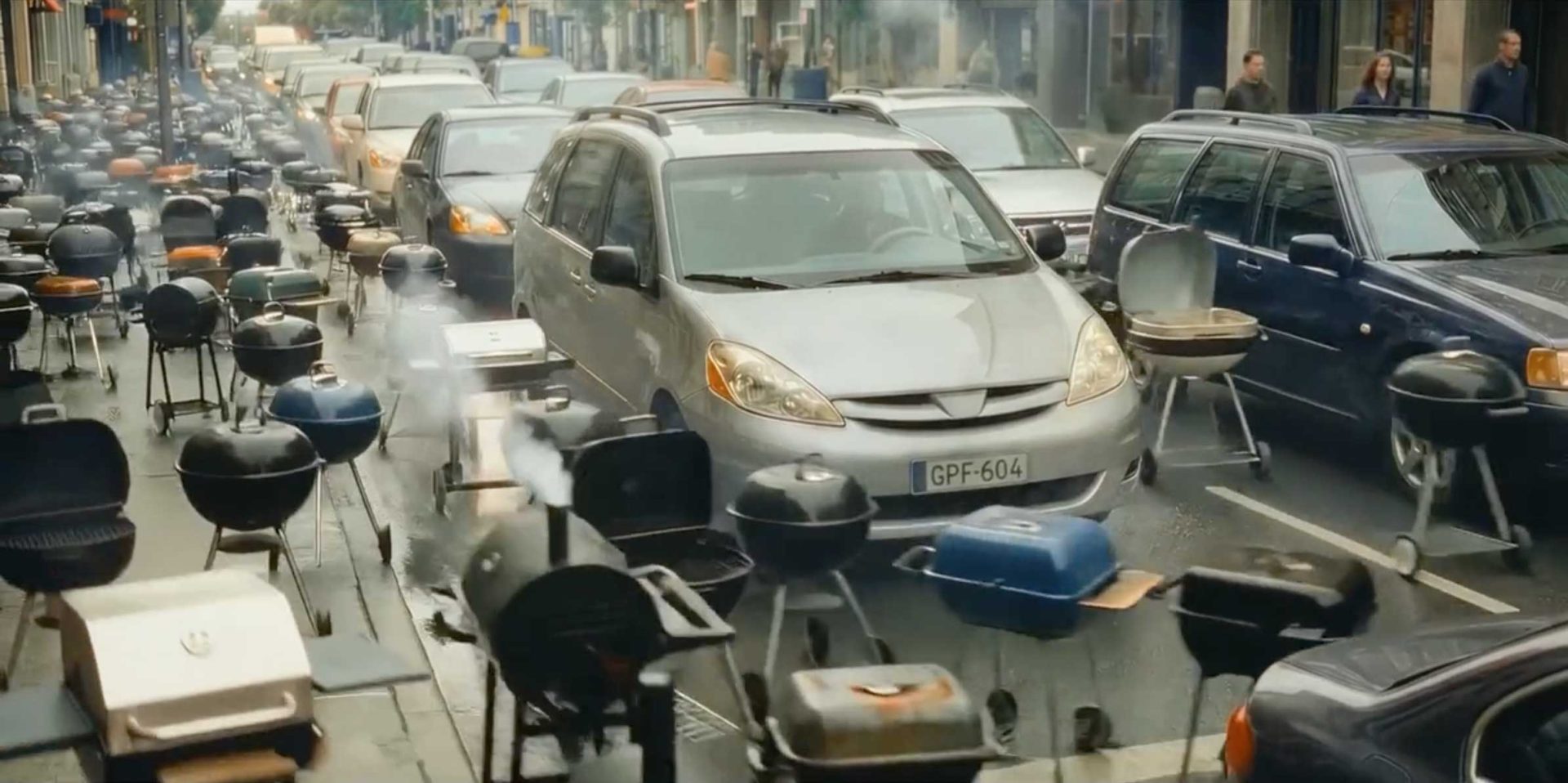 A busy city street is filled with parked cars and dozens of smoking Barbecue Piper grills, taking up the roadway where cars would normally be driving. People are seen walking on the sidewalks in the background. Frame from Stash Magazine article.