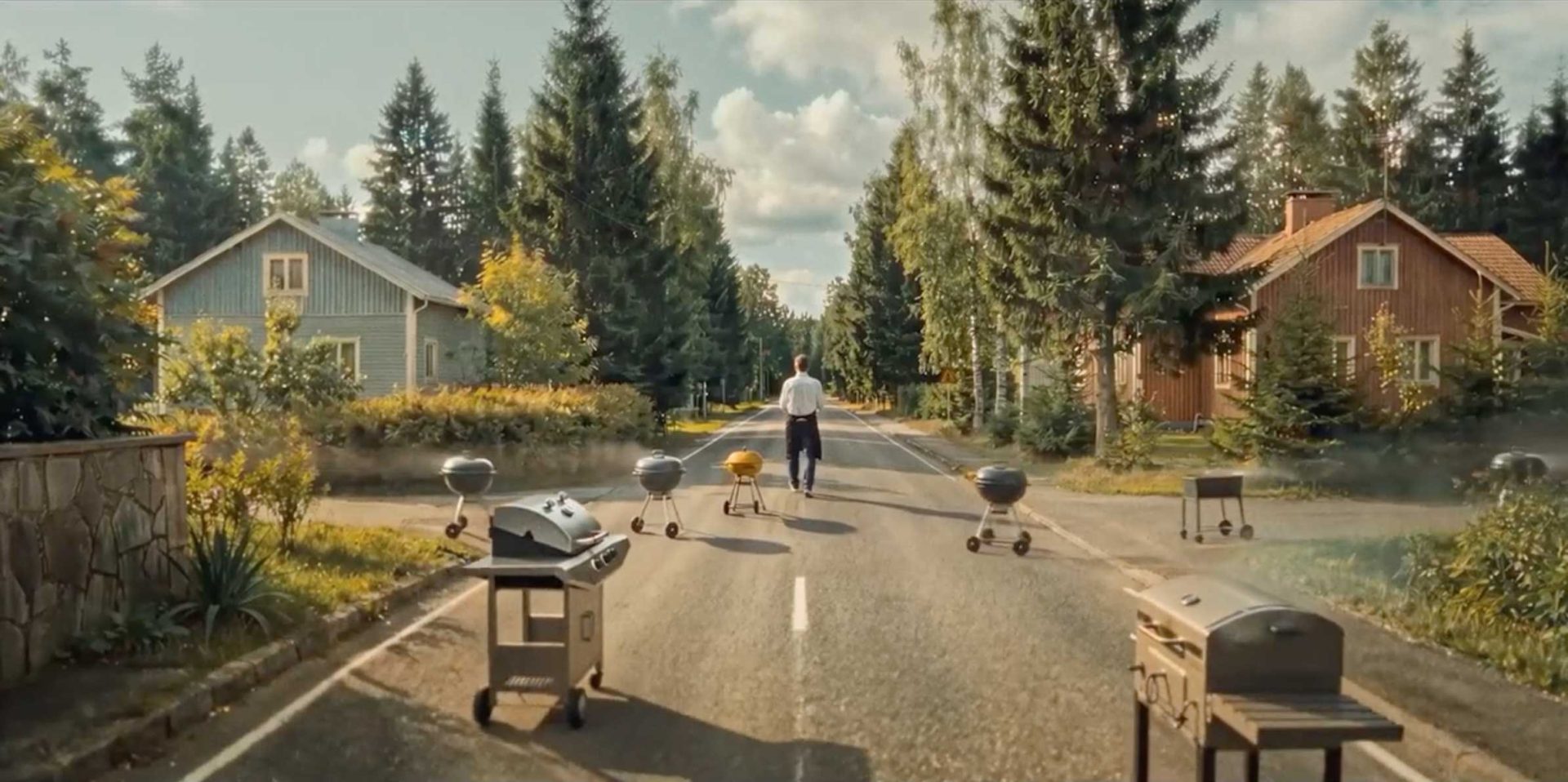 A person walks down a tree-lined street with several barbecues and grills on each side. Cozy houses with gardens line the road, where sunlight filters through the trees, creating a peaceful suburban scene reminiscent of a Lidl Finland commercial directed by Pete Riski. Frame from Stash Magazine article.