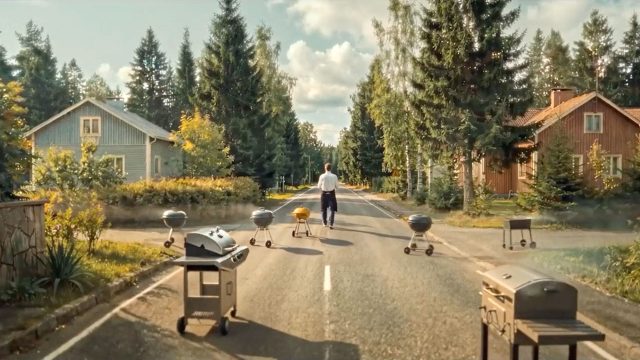 A person walks down a sunny, tree-lined street lined with barbecue grills, evoking the calm of a Pete Riski scene for Lidl Finland. Houses and tall pine trees in the background complete this peaceful Barbecue Piper setting. Frame from Stash Magazine article.