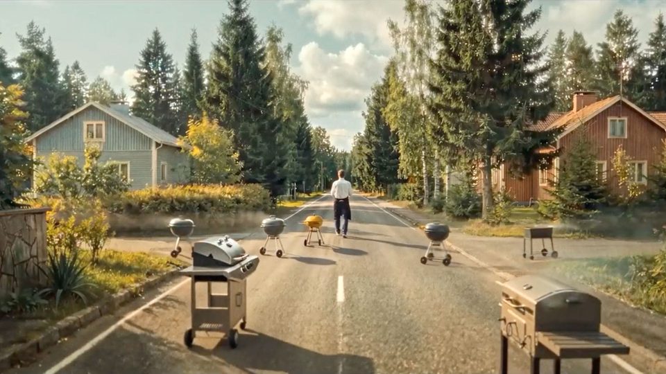 A person walks down a sunny, tree-lined street lined with barbecue grills, evoking the calm of a Pete Riski scene for Lidl Finland. Houses and tall pine trees in the background complete this peaceful Barbecue Piper setting. Frame from Stash Magazine article.