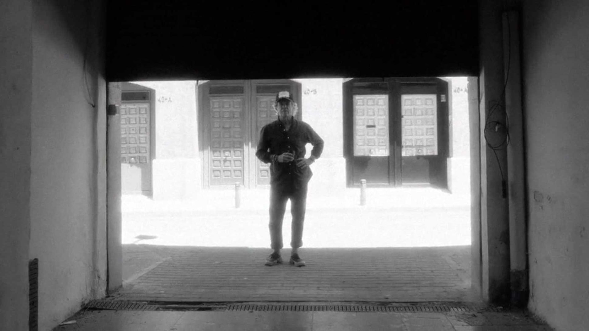 A person stands in a shadowed doorway, holding a camera, with bright sunlight and large doors visible in the background—a scene reminiscent of the bold visual storytelling seen in David Carson’s work, subtly hinting at a Brand Refresh for LOLA Global. Frame from Stash Magazine article.
