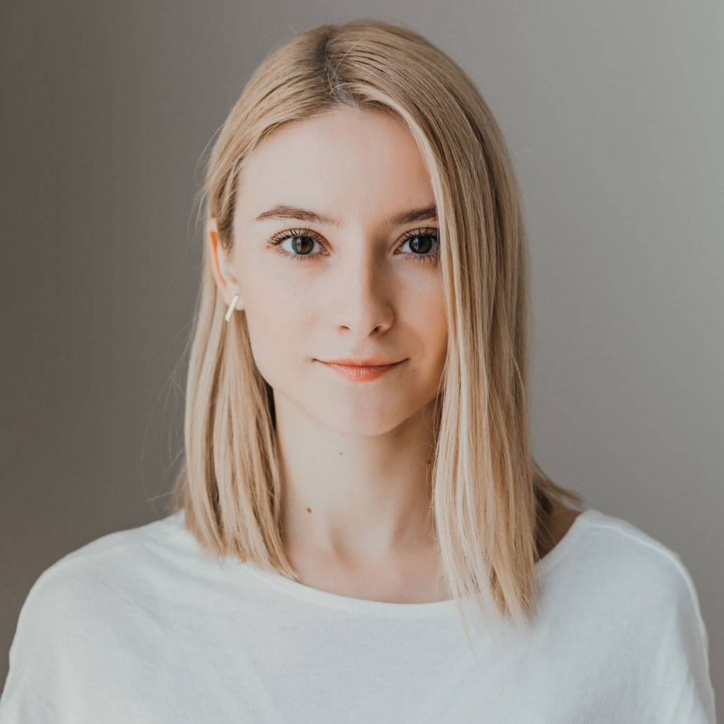 A young woman with straight, shoulder-length blonde hair and fair skin, Lidia SCARLAT wears a white top and looks directly at the camera with a neutral expression against a plain, light background. Frame from Stash Magazine article.