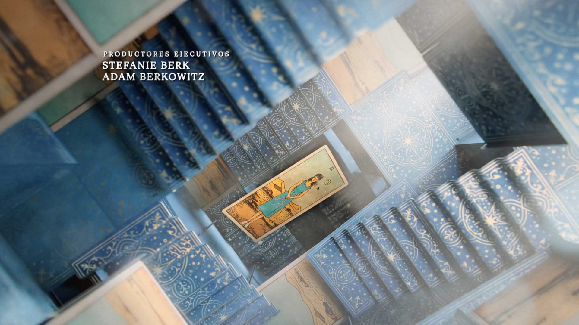 A view looking down a spiraling staircase with blue, ornate, celestial-patterned tiles, reminiscent of La Casa de los Espíritus. At the center, a framed painting of a woman in blue. Text in Spanish credits executive producers Stefanie Berk and Adam Berkowitz. Frame from Stash Magazine article.