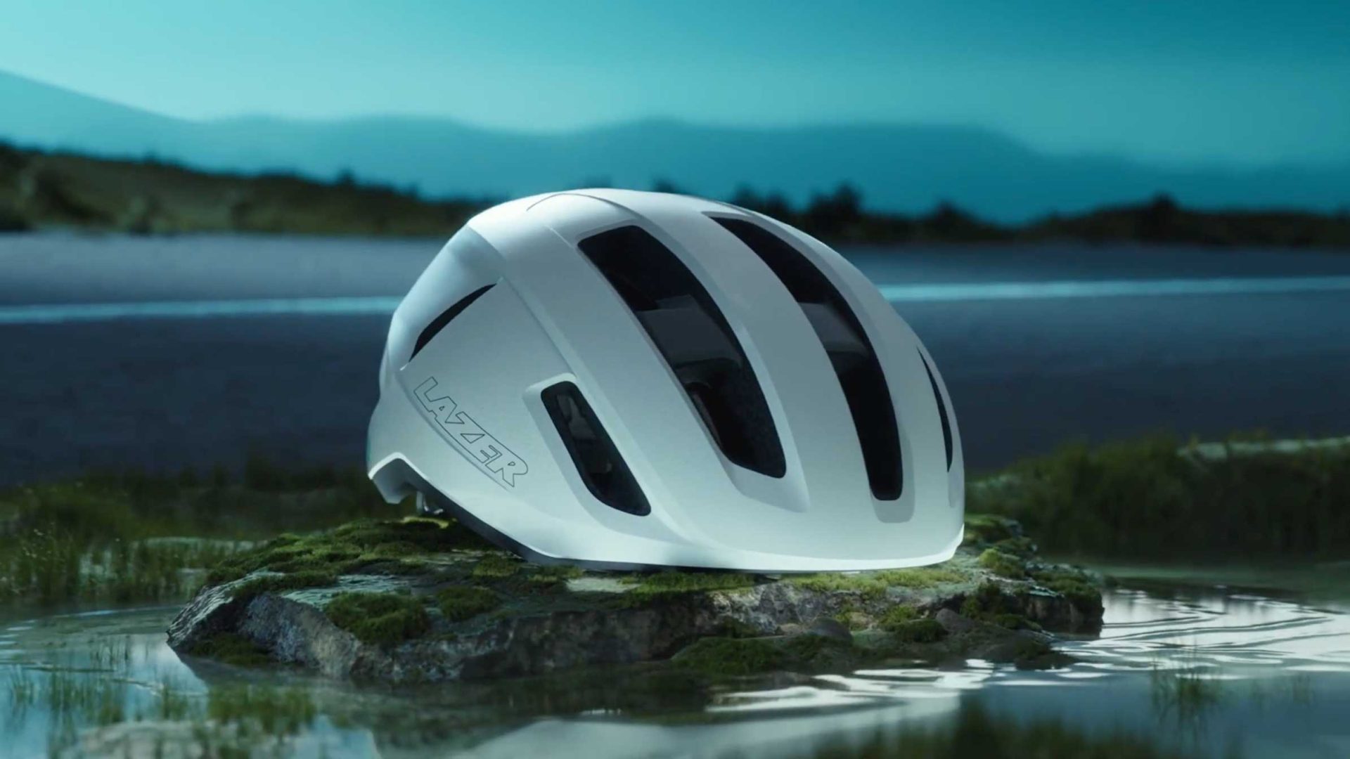 A white Lazer bicycle helmet rests on a mossy rock surrounded by shallow water, featured in Frame48's Hell & High Water, with a blurred natural landscape and mountains in the background under a blue sky. Frame from Stash Magazine article.