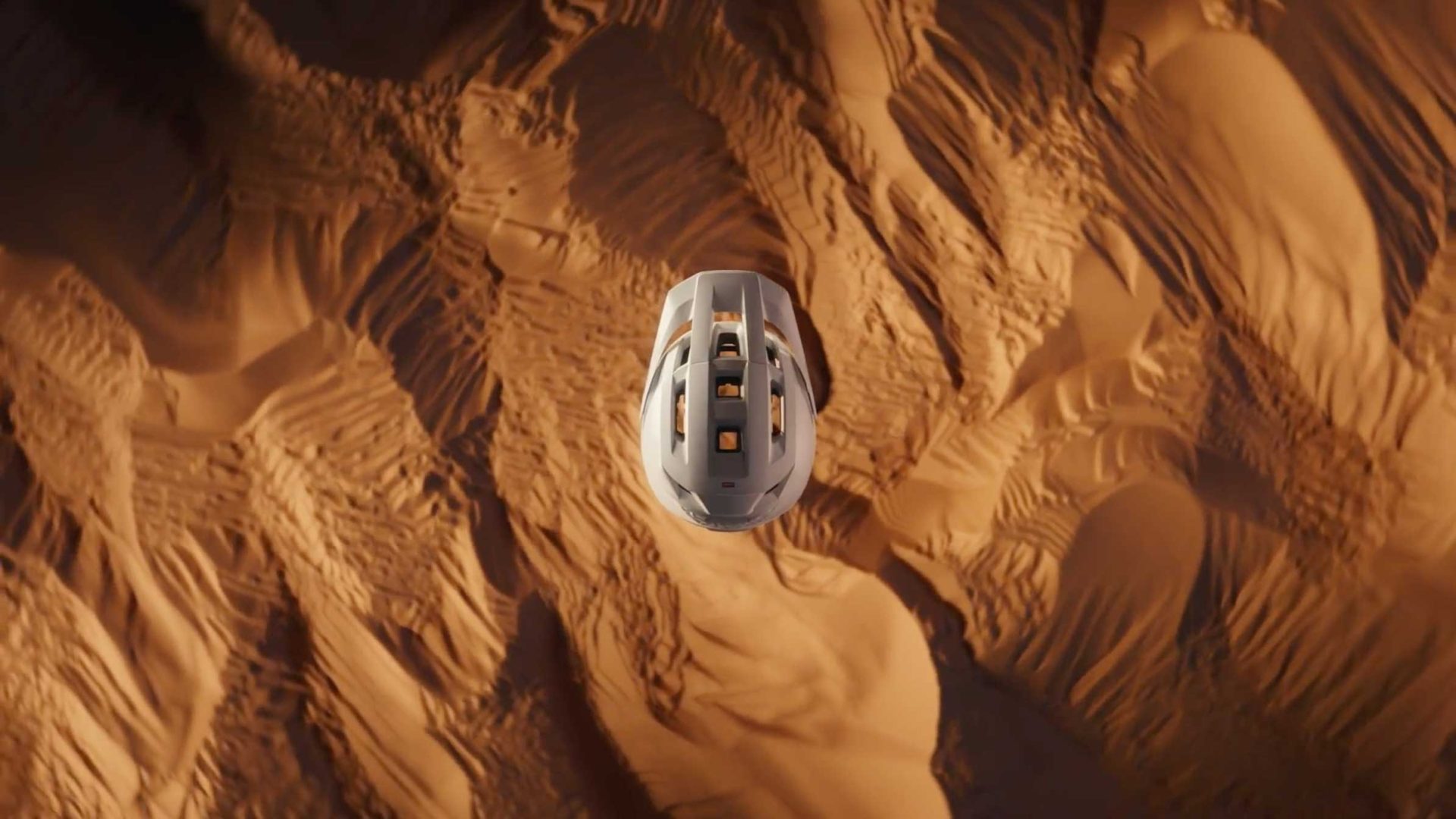 A white futuristic vehicle or pod, viewed from above, speeds over a textured, sandy desert landscape with wavy ridges—like a scene from Hell & High Water by Frame48. Frame from Stash Magazine article.
