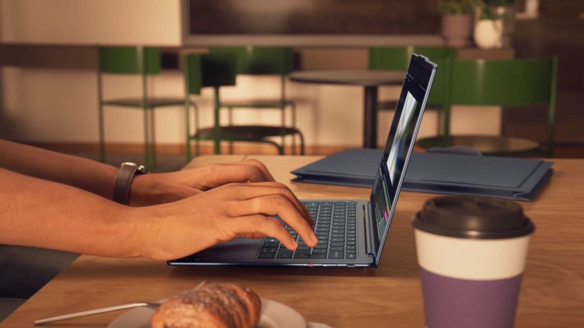 A person types on a Lenovo laptop at a wooden table in a cafe, with a coffee cup, a pastry, and folders nearby. Green chairs and the blurred background suggest a relaxed, casual atmosphere captured in the inviting style of Frame48. Frame from Stash Magazine article.