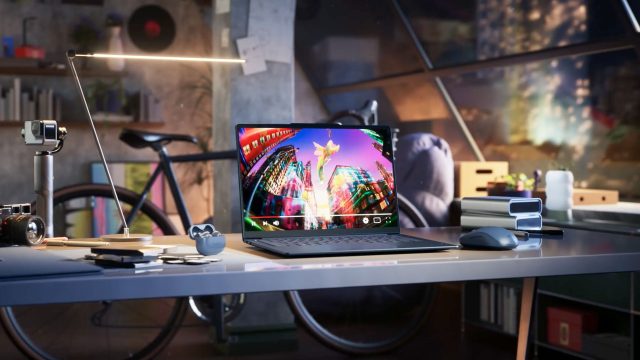 A modern workspace featuring a Lenovo laptop on a glass desk, displaying colorful city lights. Nearby are headphones, a mouse, a lamp, a cup, and a bicycle, with large windows showing an evening cityscape—capturing the creative spirit of Frame48. Frame from Stash Magazine article.
