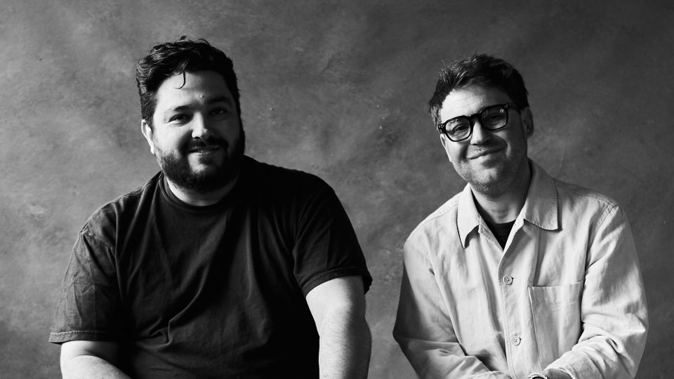 Two men sitting side by side against a plain, textured backdrop—one wears a dark t-shirt and has a beard; the other wears glasses and a light button-up shirt. Both are smiling, exuding warmth as they mark the beginning of Little Bird Signs CARAVAN Signs for East Coast Representation. The photo is in black and white. Frame from Stash Magazine article.