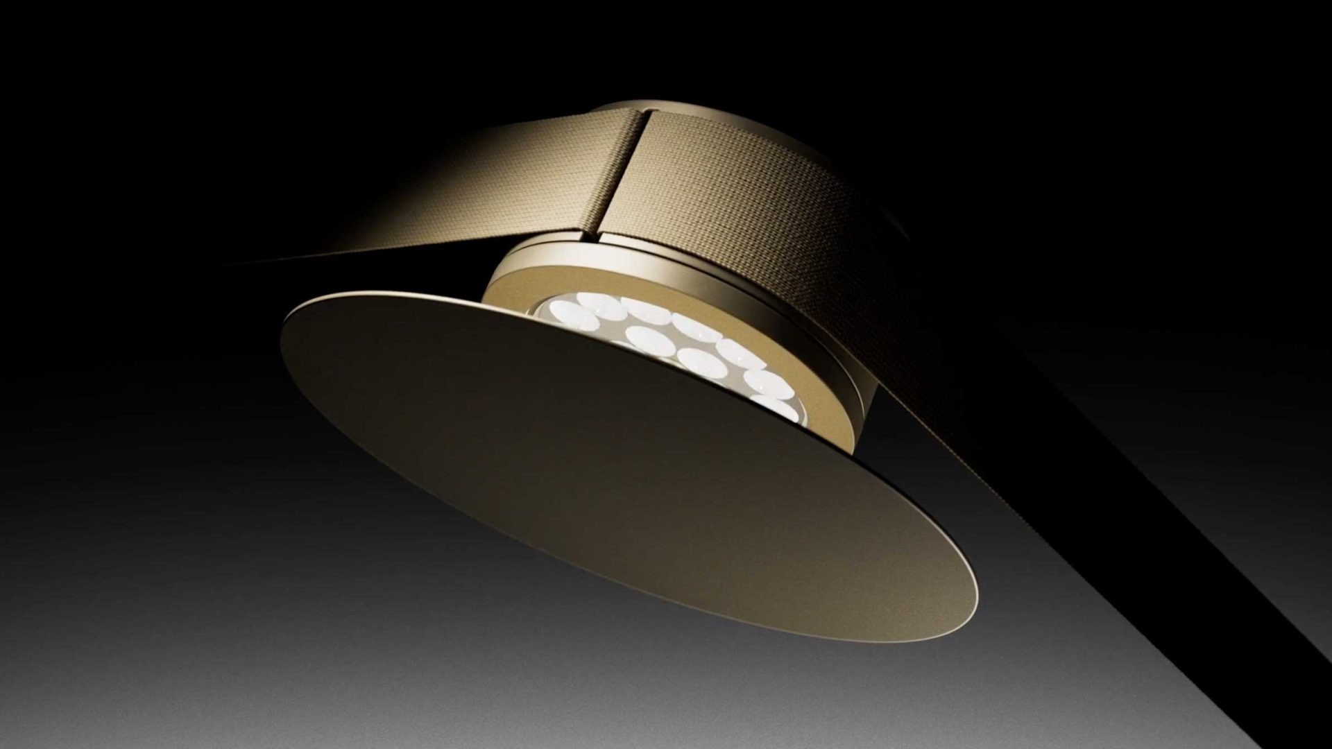 Close-up of a modern, gold-colored LED ceiling light fixture by Lodes with a circular design, set against a dark, gradient background. The light is angled, highlighting its sleek, textured metallic surface—a signature of MAP lighting. Frame from Stash Magazine article.