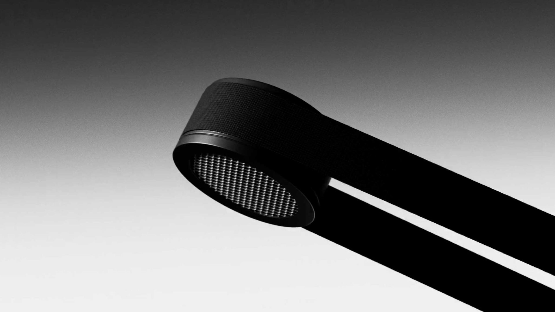 A minimalistic black modern lamp with a cylindrical head and mesh-like grill, this Lodes MAP lighting product is attached to two flat horizontal supports and shown against a smooth gradient gray background. Frame from Stash Magazine article.
