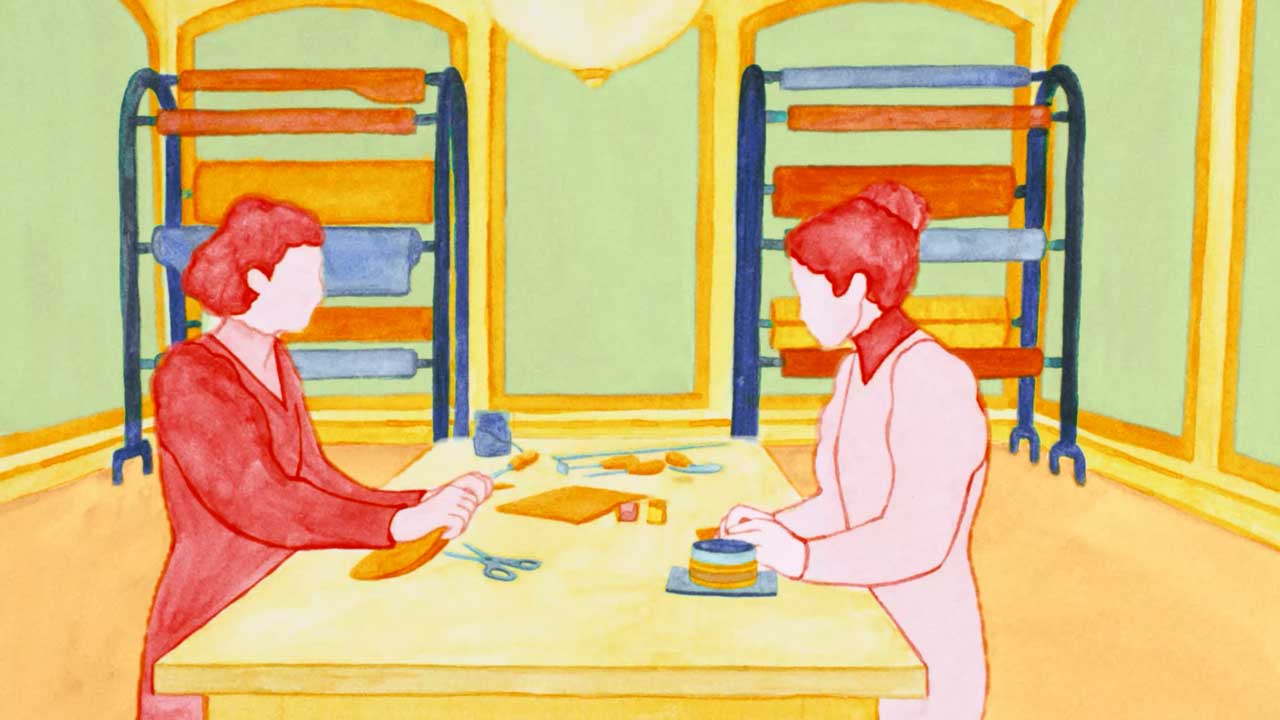 Two people sit across from each other at a table, working with fabric and sewing supplies. Behind them, shelves hold colorful rolls of material. The scene evokes the artistry seen in the Loewe 180th anniversary brand film by Isabel Garrett & Blinkink. Frame from Stash Magazine article.