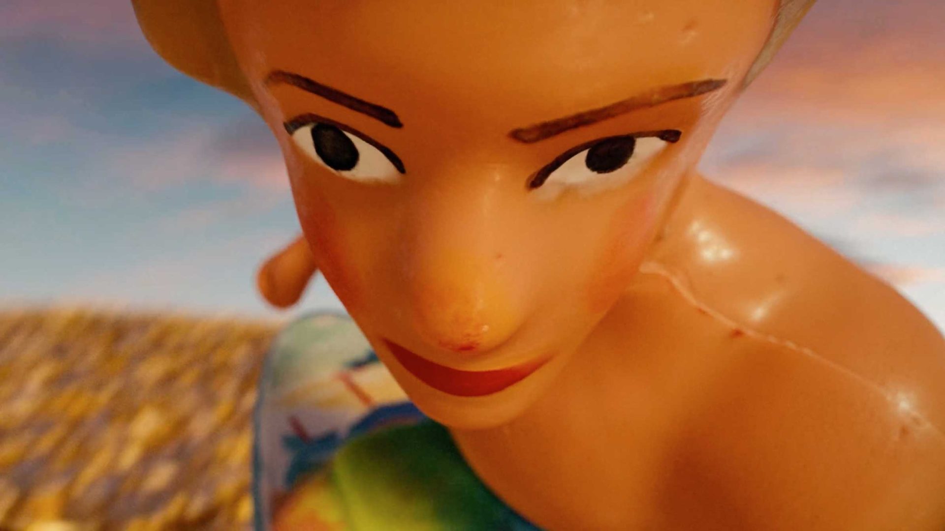 Close-up of a plastic doll with painted facial features and blonde hair, looking directly at the camera. The background is blurry, showing a blue sky and a yellow surface, possibly sand—reminiscent of a whimsical spec spot for Crumb, the fintech brand. Frame from Stash Magazine article.