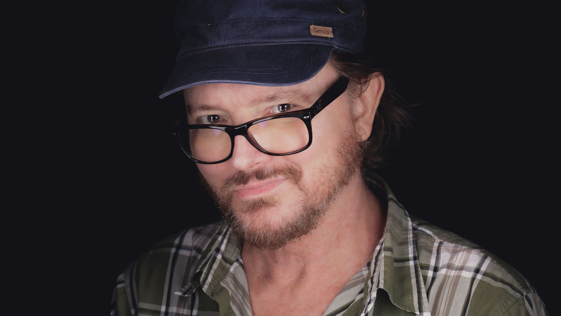 A person with a short beard and glasses, reminiscent of the professionals leading creative teams, wears a blue cap and plaid shirt, looking into the camera with a slight smile against a dark background—echoing the recent news as Lux-Aeterna appoints Richard Stay as Compositing Supervisor. Frame from Stash Magazine article.