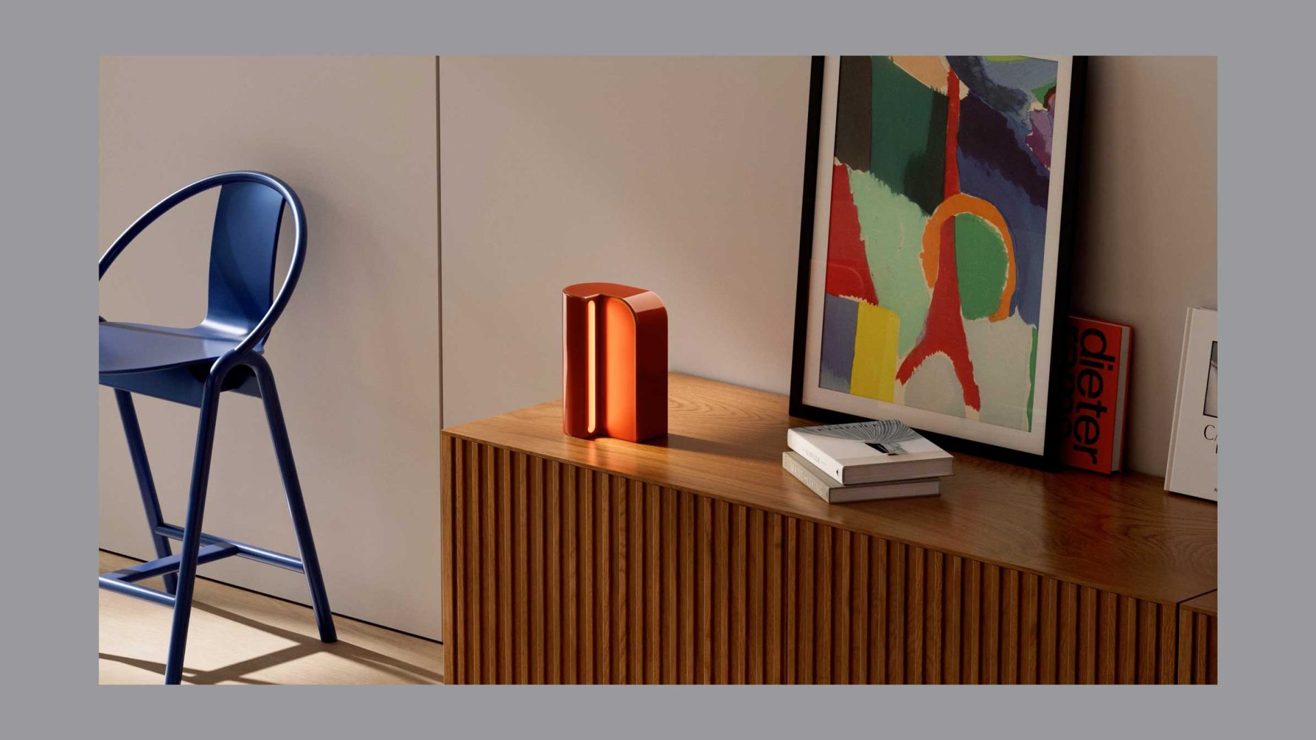 A modern interior features a blue chair, a wooden sideboard by MADE, an abstract framed artwork, books, and a small, orange sculptural object on a wood cabinet against a neutral wall. Frame from Stash Magazine article.