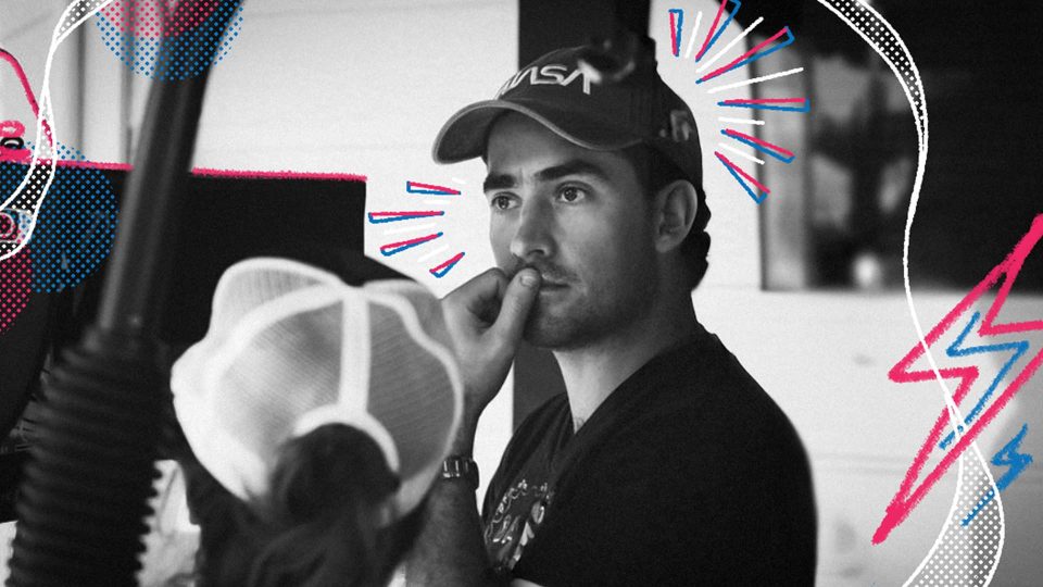 A person wearing a NASA cap looks ahead as someone applies makeup to their face. The image is black and white with colorful graphic doodles and lines added around the subjects.