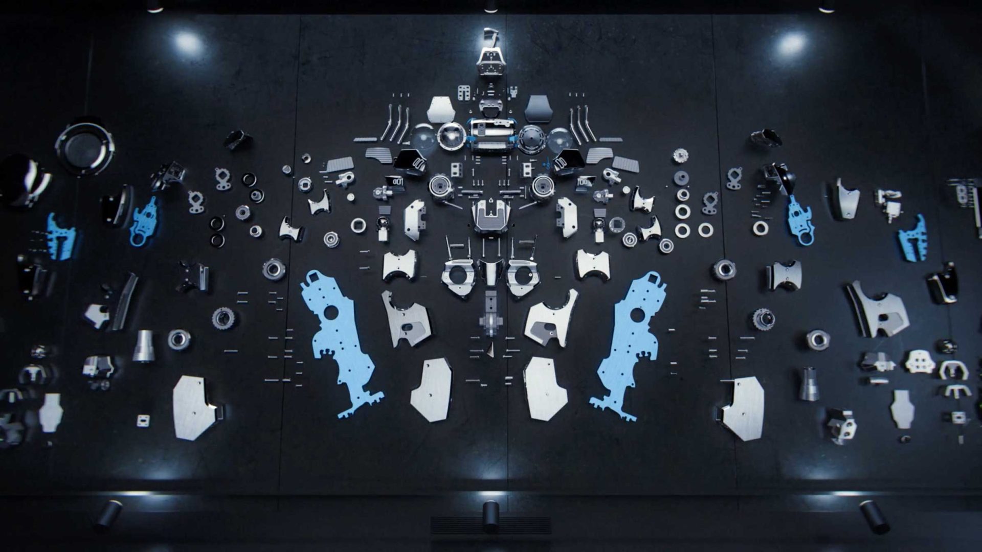 A symmetrical arrangement of various mechanical parts and components, including blue plates, metallic gears, bolts, and brackets—reminiscent of MB&F's HM12 design—laid out on a dark surface and illuminated by overhead lights. Frame from Stash Magazine article.