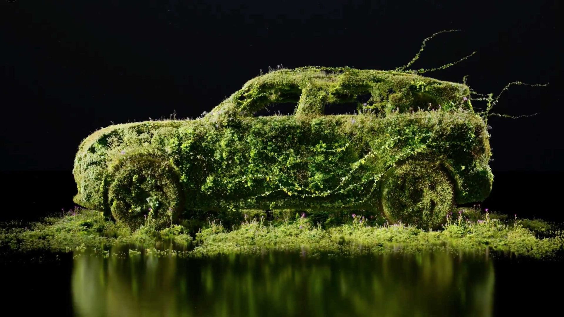 A car completely covered in green moss and plants sits against a black background, with its reflection visible on a shiny surface below—an imaginative nod to MINI Icon Drops and Special Editions. Frame from Stash Magazine article.