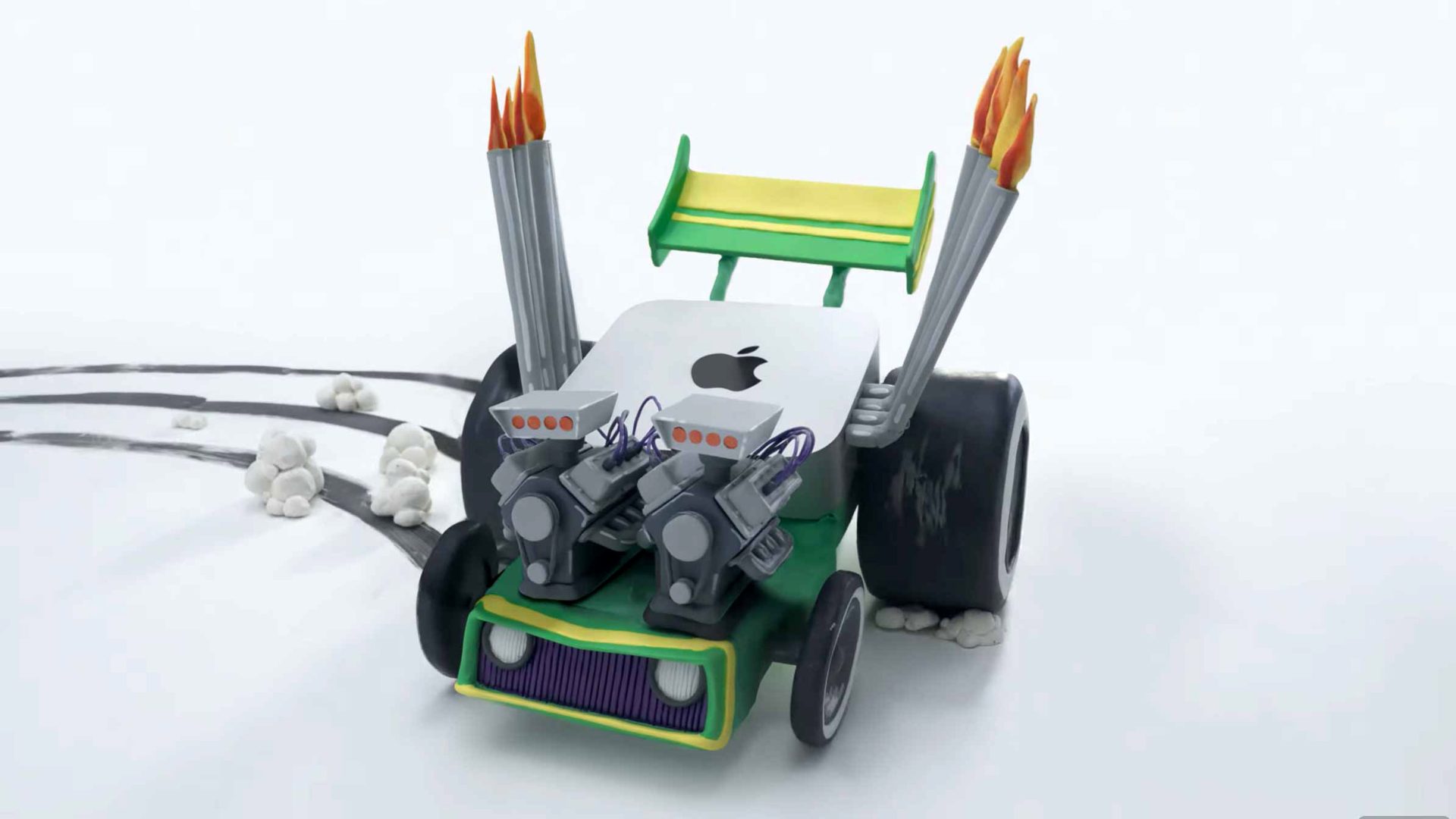 A toy-like race car with an Apple logo, large rear tires, twin exhaust pipes with flames, engine parts on the hood, and tire skid marks on a white surface. Frame from Stash Magazine article.