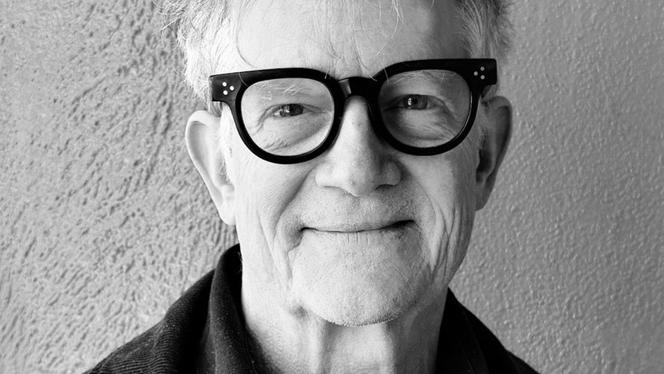 Older man with light hair, large dark glasses, and a slight smile stands in front of a textured wall. The image is in black and white and shows him from the shoulders up, capturing the thoughtful presence of Alex McDowell, an innovator known for his contributions to New Narrative Design. Frame from Stash Magazine article.
