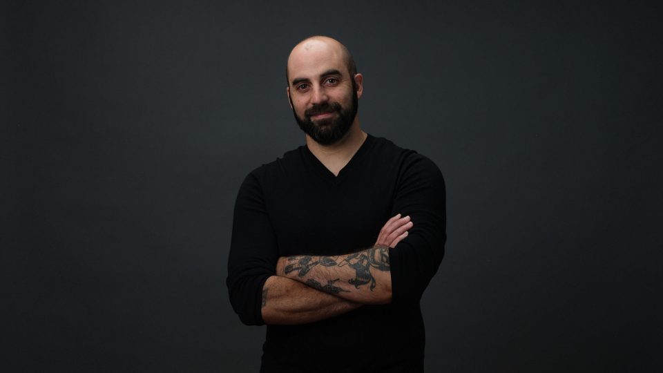 A man with a beard and tattoos stands against a dark background, wearing a black long-sleeve shirt and smiling slightly with his arms crossed—exuding the confident presence of Marc Fusco, Head of Production. Frame from Stash Magazine article.