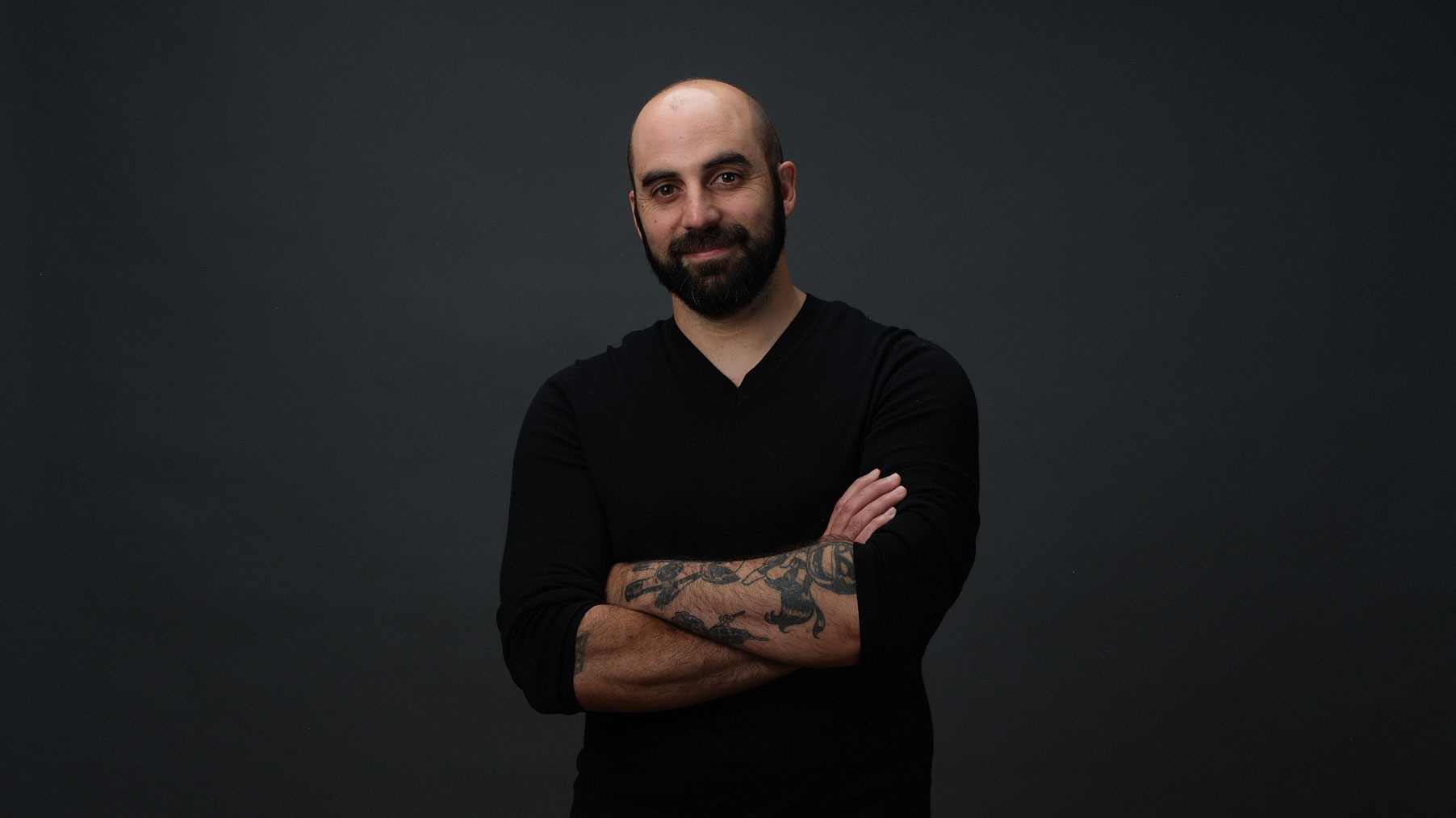 A man with a beard and tattoos stands against a dark background, wearing a black long-sleeve shirt and smiling slightly with his arms crossed—exuding the confident presence of Marc Fusco, Head of Production. Frame from Stash Magazine article.