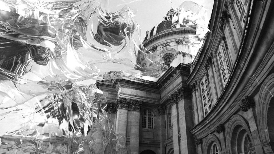 A flowing, translucent, abstract sculpture stands in front of a grand historic building with columns and a domed roof, seen in black and white, inspired by Martynas Genkovas’s visionary style. Frame from Stash Magazine article.