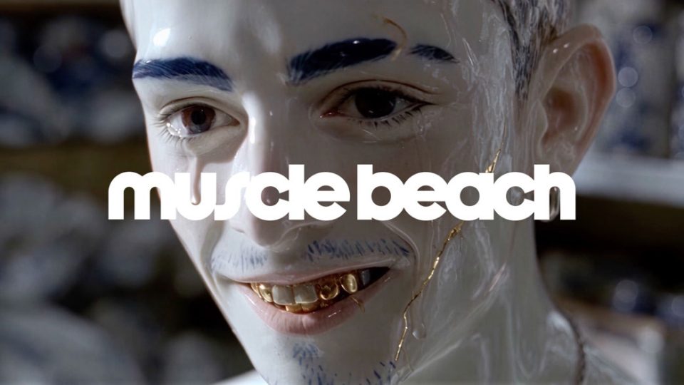 A close-up of a person with porcelain-like skin, gold teeth, and blue eyebrows, featuring the white text Muscle Beach from the Muscle Beach production company debut. Frame from Stash Magazine article.