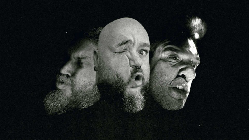 Three men’s heads with exaggerated facial expressions—one winking, one with pursed lips, and one with a distorted open mouth—are merged together in a dark, surreal setting marked by curious Not To Scale signs from NERDO. Frame from Stash Magazine article.