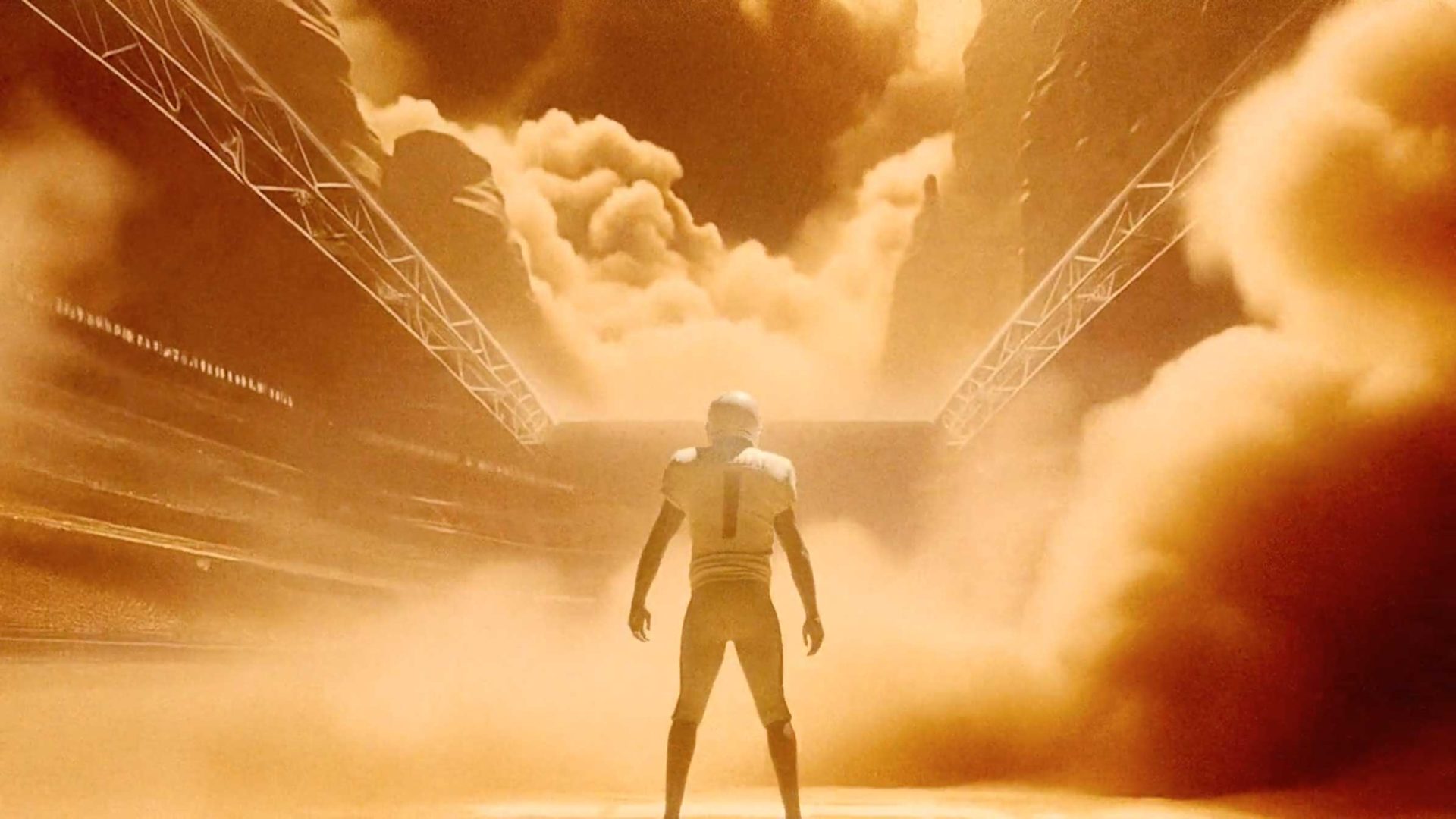 A football player stands alone on a field, facing away, as dramatic orange-brown clouds of dust and light—echoing the intensity of the NFL and Nike Rivalries Uniforms release film by Builders Club—fill the stadium, creating a surreal atmosphere. Frame from Stash Magazine article.