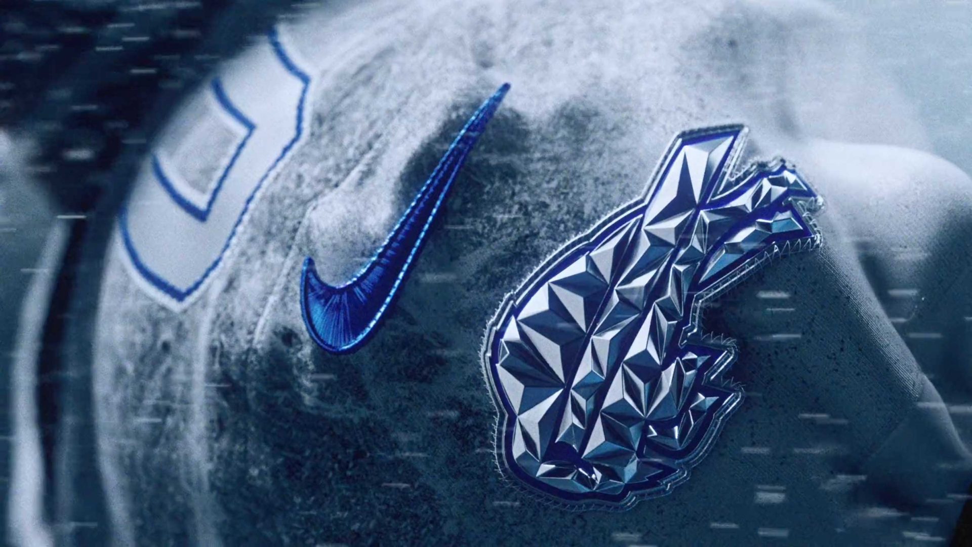 A close-up of a sports jersey featuring a blue Nike swoosh and a shiny, faceted, metallic panther logo, with snow or frost covering the fabric. The NFL number 0 is partially visible in the background. Frame from Stash Magazine article.