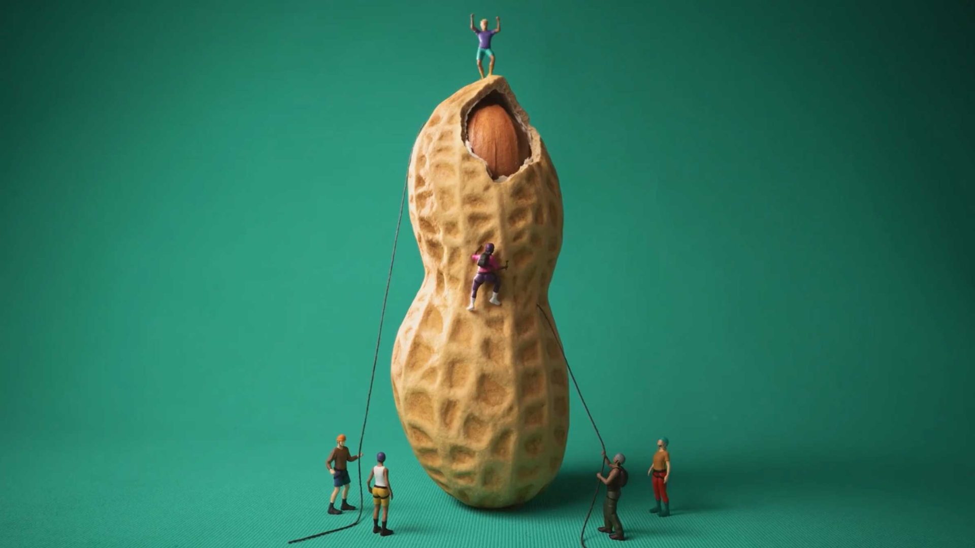 Five miniature figurines climb and cheer around a large peanut shell against a solid green background, with one figure celebrating on top—an imaginative scene inspired by Anthony Farquhar-Smith and his playful take on peanuts. Frame from Stash Magazine article.