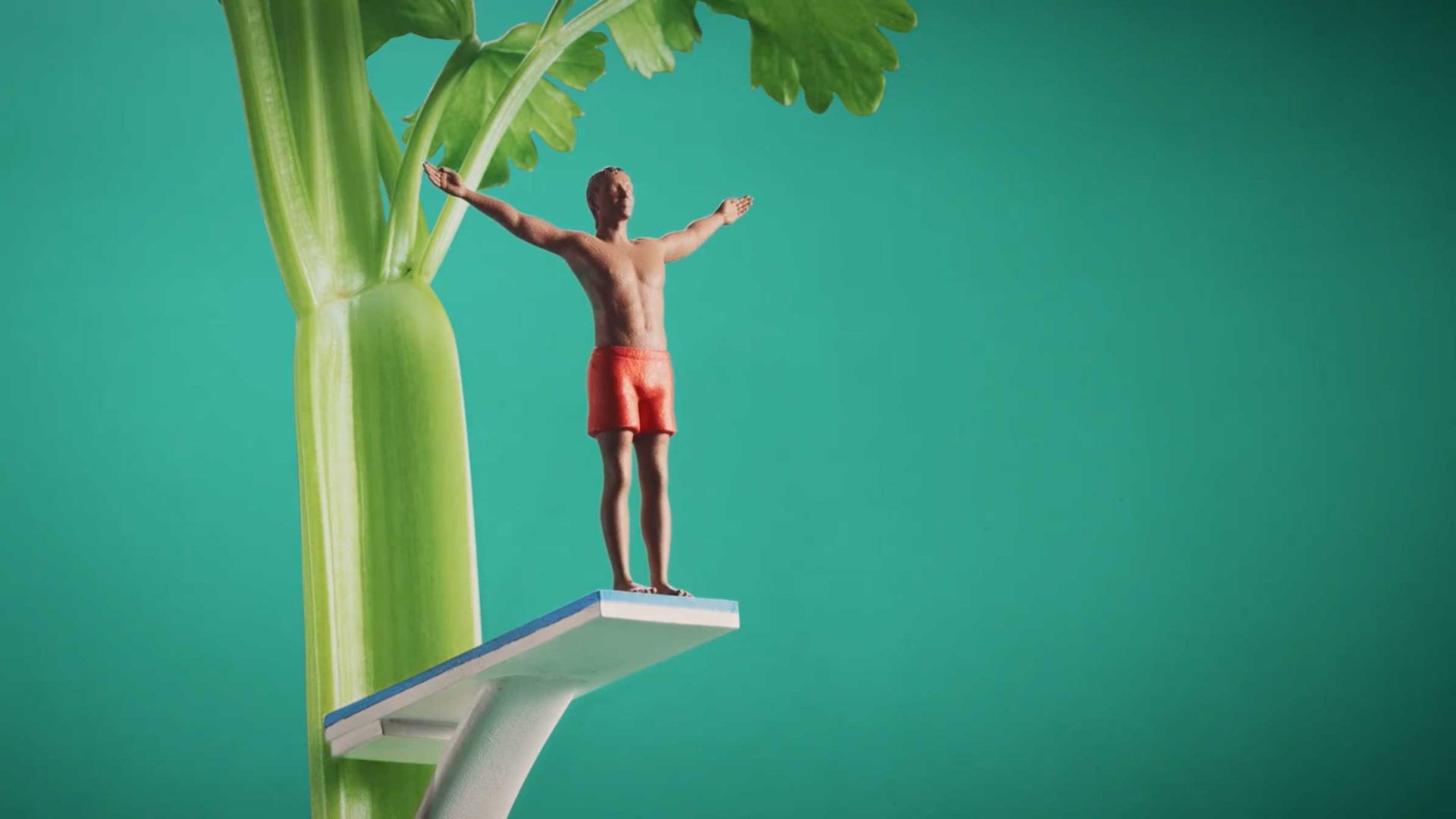 A small figurine of a man in red swim trunks stands with arms outstretched on a diving board attached to a large stalk of celery, against a green background—definitely not to scale. Frame from Stash Magazine article.
