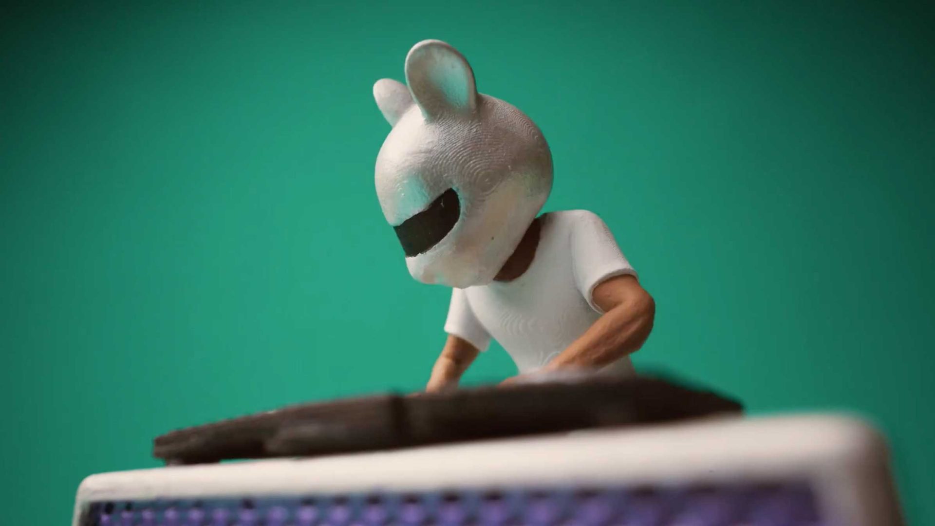 A figurine with a white mouse-like helmet and a white shirt stands behind a DJ table, appearing to mix music, against a green background—reminiscent of Anthony Farquhar-Smith’s playful style in films like Peanuts. Frame from Stash Magazine article.