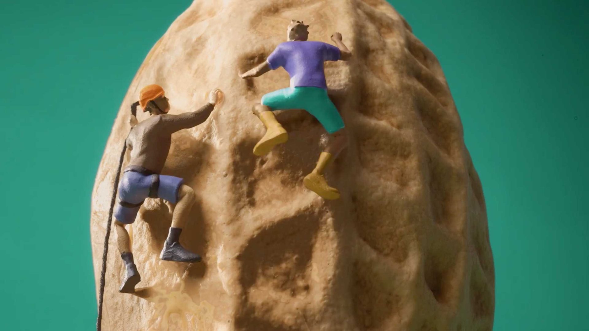 Two miniature figurines in climbing gear appear to scale the surface of a large peanut shell against a green background, cleverly creating the illusion of rock climbing on peanuts—a whimsical scene reminiscent of Anthony Farquhar-Smith’s playful style. Frame from Stash Magazine article.