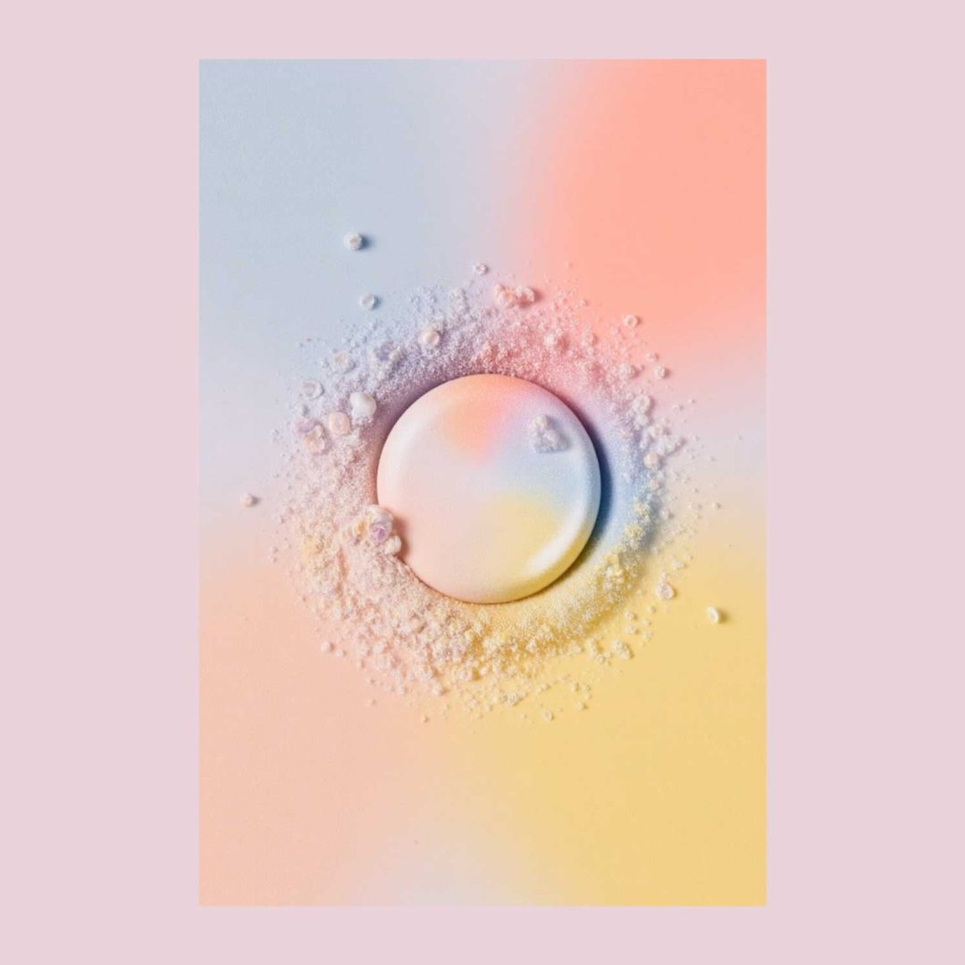 A round tablet surrounded by powder sits on a pastel gradient background of pink, orange, and yellow hues, creating a soft and dreamy effect reminiscent of Vitor Teixeira's personal short film "Unfurl. Frame from Stash Magazine article.