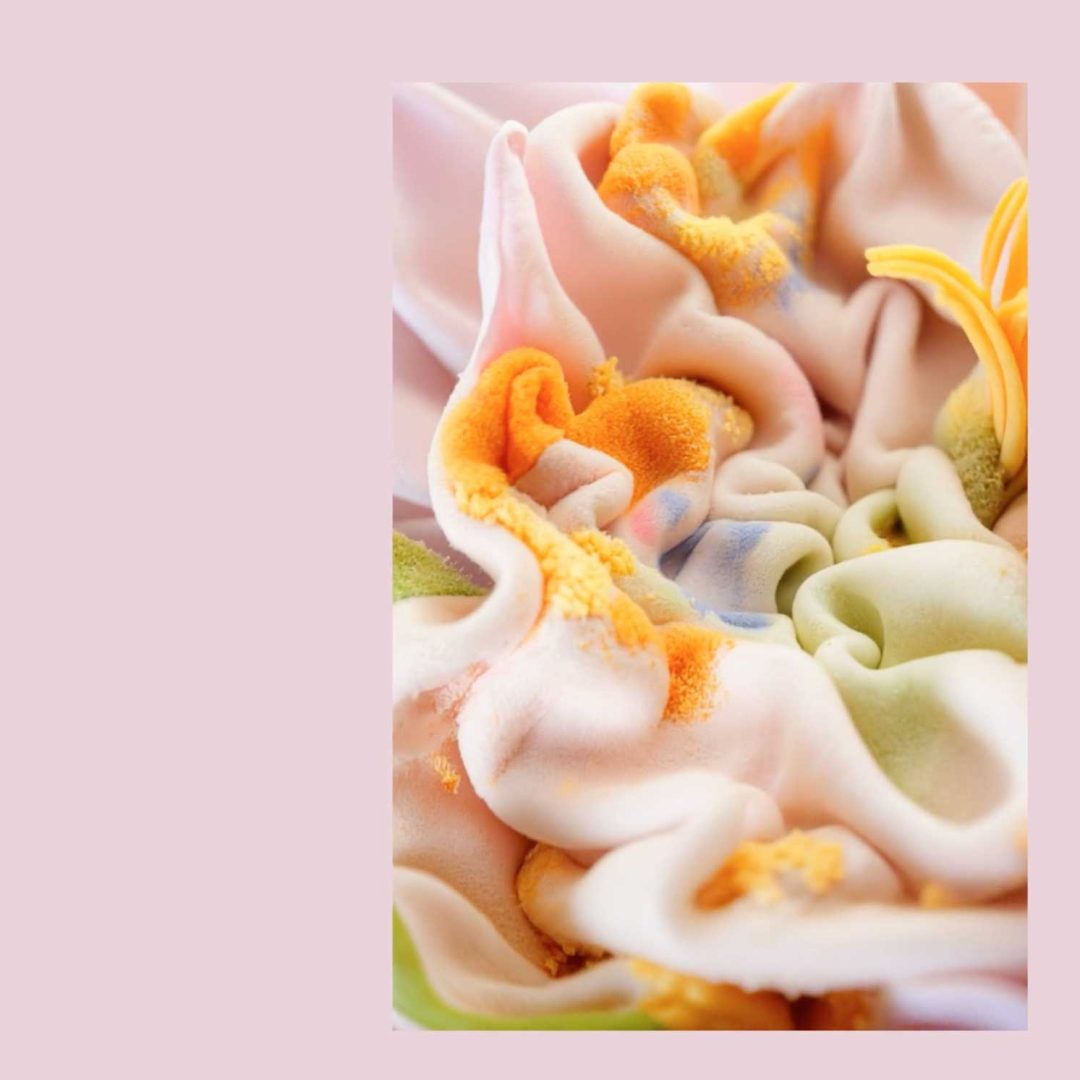 Close-up of pastel-colored, ruffled fabric with orange, yellow, and green accents against a pale pink background, echoing the abstract flower petals and flowing folds featured in Vitor Teixeira's short film "Unfurl. Frame from Stash Magazine article.