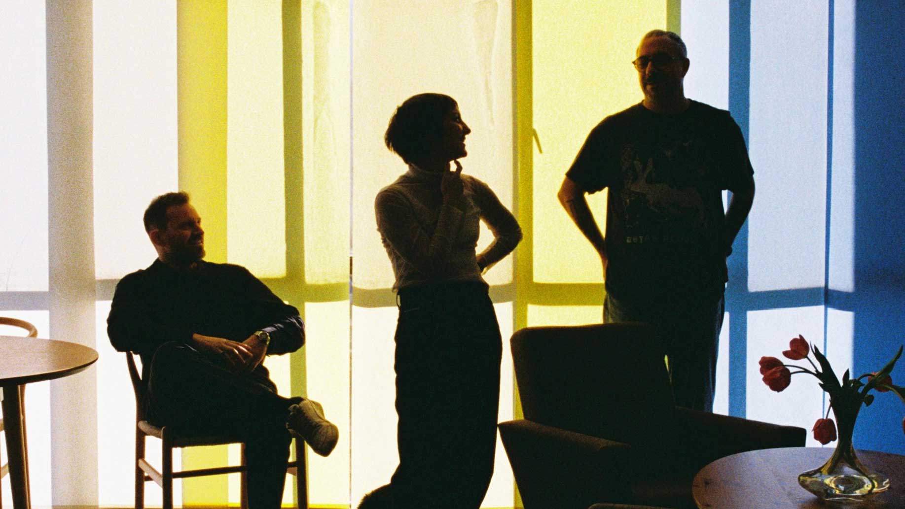 Three people are silhouetted against a large, softly lit window with yellow and blue panels. A vase with tulips sits on a table in the foreground—a scene inspired by the Niceshit studio rebrand with a custom font aesthetic. Frame from Stash Magazine article.