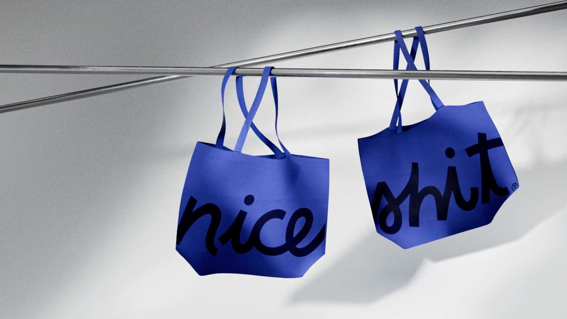 Two blue tote bags hang on metal rods. One bag has the word “nice” in bold black script, and the other has the word “shit” in the same style. The bags cast soft shadows on a white background. Frame from Stash Magazine article.