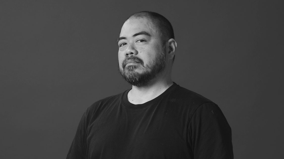 A man with a beard and mustache, identified as Nico-Puertollano-Versus-ECD, wears a black T-shirt and stands against a plain, dark background, looking slightly to the side with a neutral expression in this black-and-white image. Frame from Stash Magazine article.