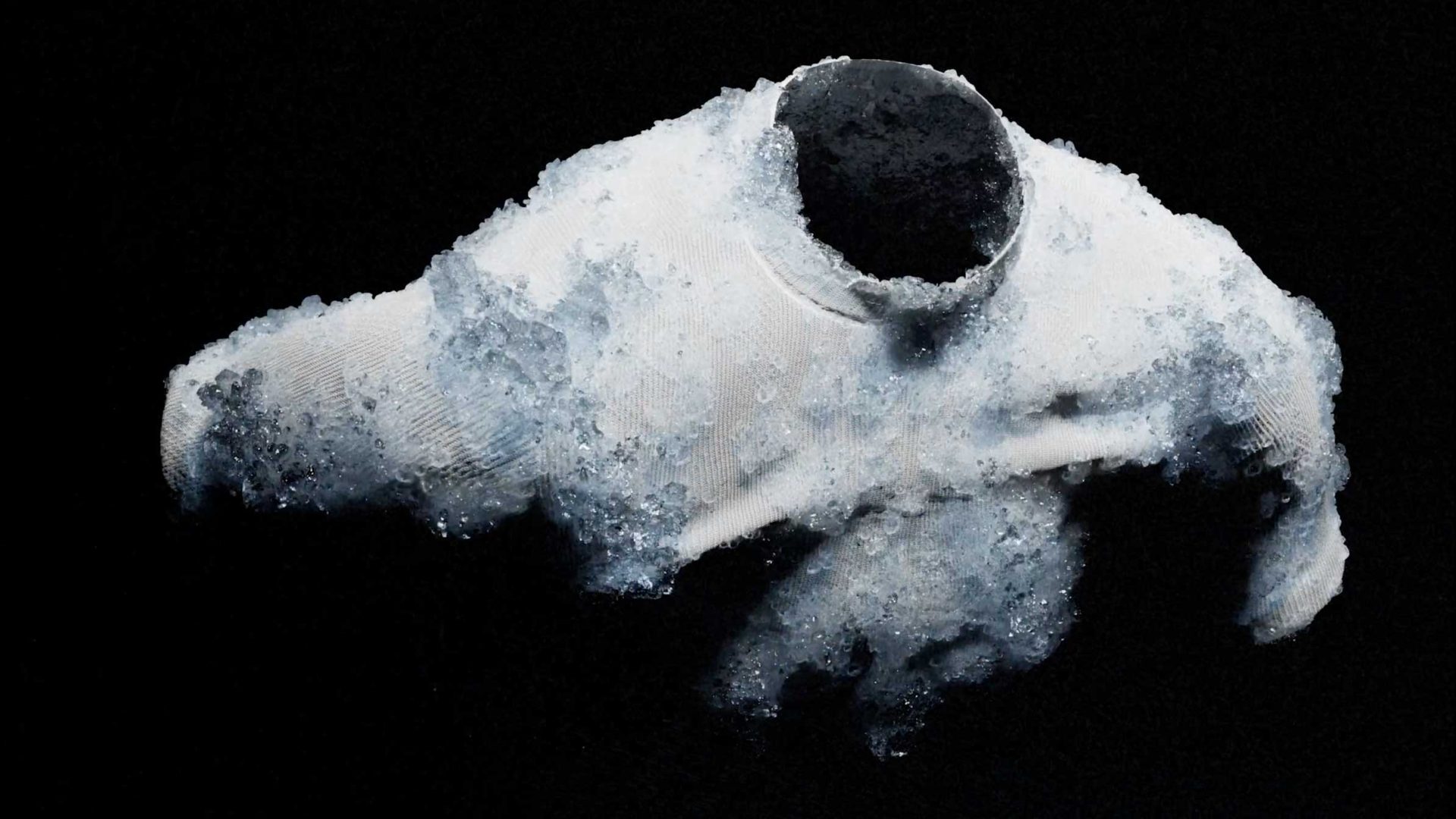A white sweater, reminiscent of Nike SP26 style, appears hollow and worn, with ice or frost covering parts of it against a black background. The garment seems to be disintegrating or melting into icy crystals. Frame from Stash Magazine article.