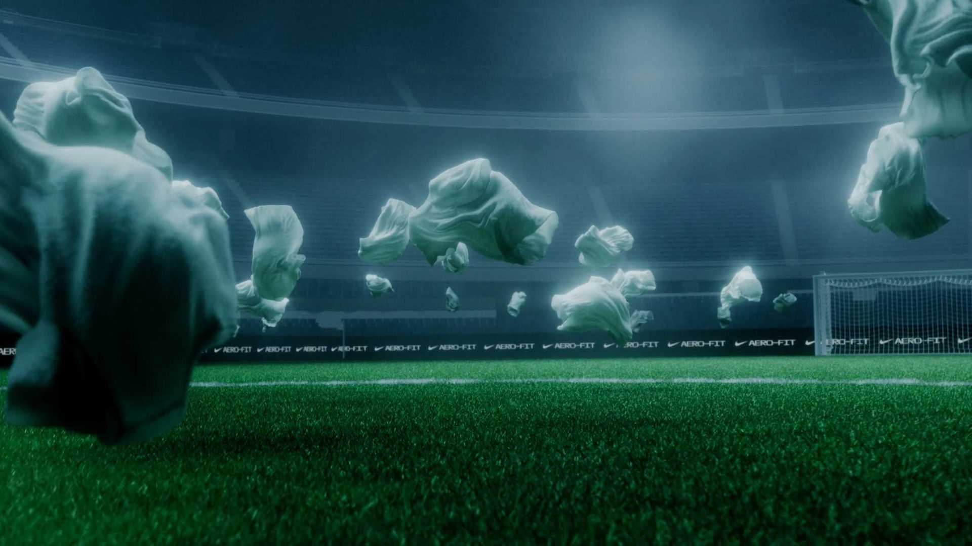 Loose white shirts by Nike SP26 float mid-air above a green soccer field inside a large, empty stadium, bright lights illuminating the haze and a goal in the background—a surreal scene crafted by Raj Davsi and FutureDeluxe. Frame from Stash Magazine article.