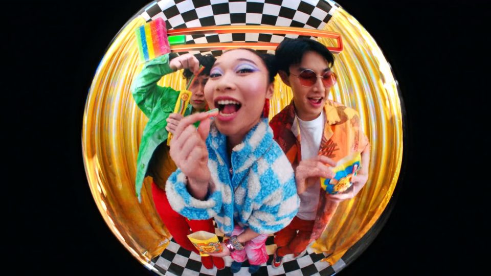 Three people in colorful Jovardi clothing smile while eating snacks, captured through a fisheye lens against a checkered and gold-curtain background. Frame from Stash Magazine article.
