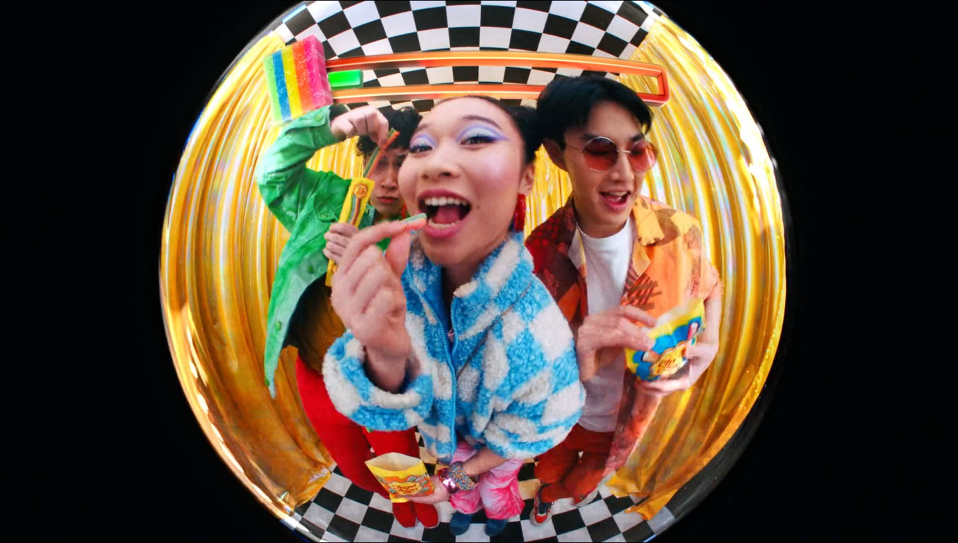 Three people in colorful Jovardi clothing smile while eating snacks, captured through a fisheye lens against a checkered and gold-curtain background. Frame from Stash Magazine article.