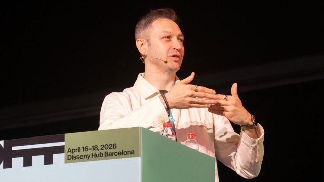 A man wearing a headset microphone gestures while speaking on stage at a podium labeled April 16-18, 2026 Disseny Hub Barcelona during the OFFF 2026 x Stash interview with Onar Senturk. A water bottle and badge are visible on the podium. Frame from Stash Magazine article.