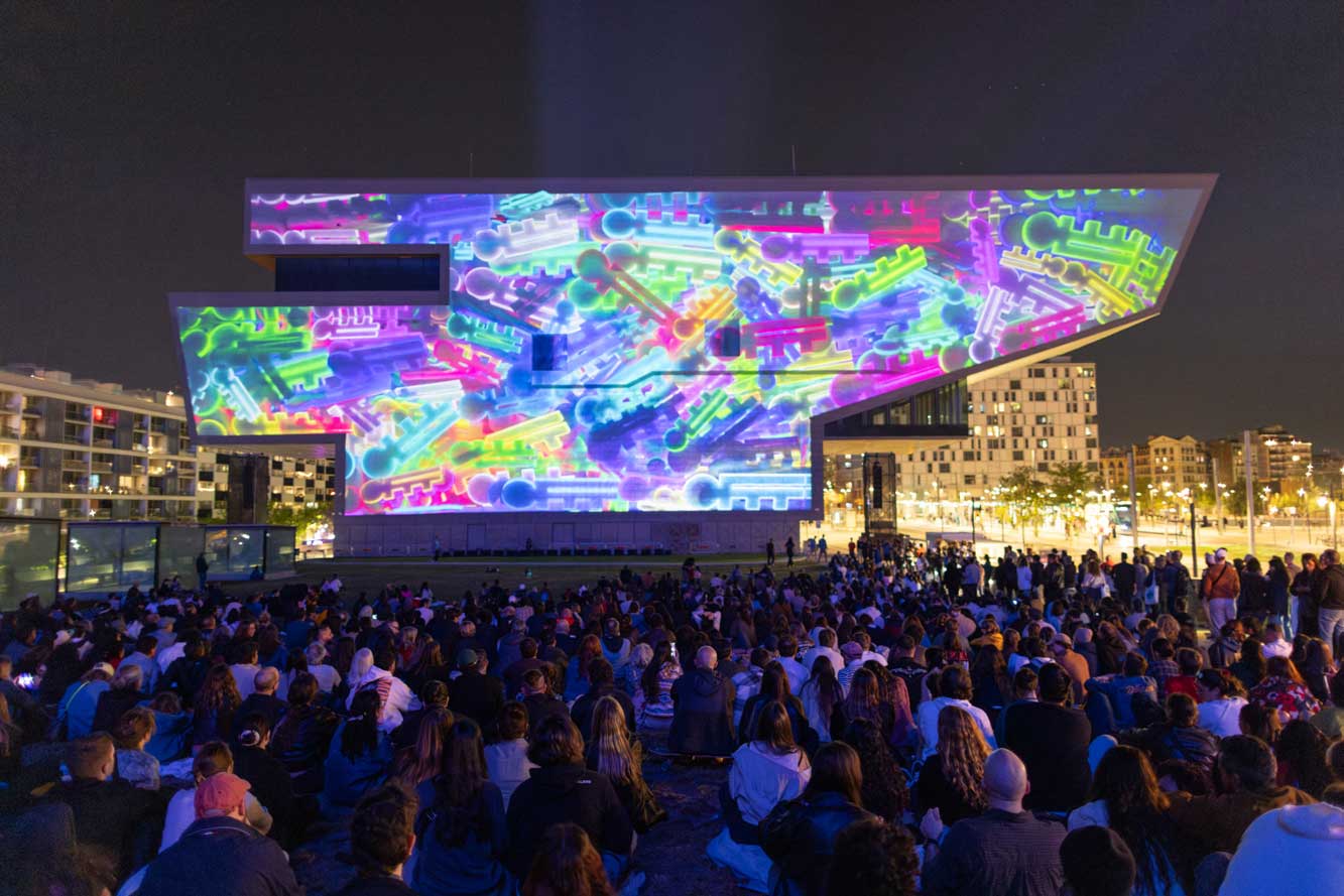 A large crowd sits outdoors at night, watching the OFFF Barcelona 2026 Main Titles—a colorful, abstract digital light projection—illuminate the facade of a modern, angular building surrounded by city lights. Frame from Stash Magazine article.