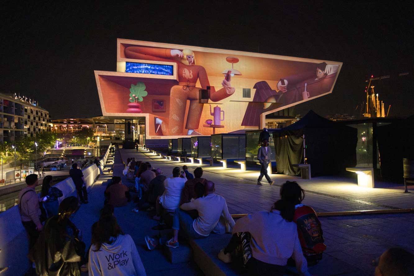People sit outdoors at night watching the Main Titles for OFFF Barcelona 2026, as a large building facade is illuminated with colorful, abstract artwork featuring human figures and objects—celebrating the event’s Rebellious DNA. The city skyline glows in the background. Frame from Stash Magazine article.