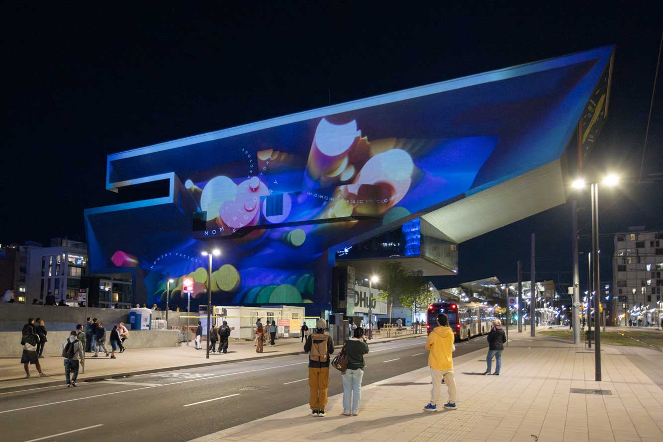 A modern, angular building lit up at night with colorful, abstract light projections for the OFFF Barcelona 2026 Main Titles, while people walk and stand on the well-lit street and sidewalk below. Frame from Stash Magazine article.