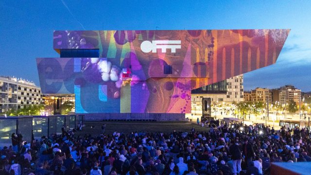 A large crowd gathers at dusk in front of a modern, angular building illuminated with colorful digital projections and the words OFFF Barcelona 2026 Main Titles displayed, during a vibrant public festival. Frame from Stash Magazine article.