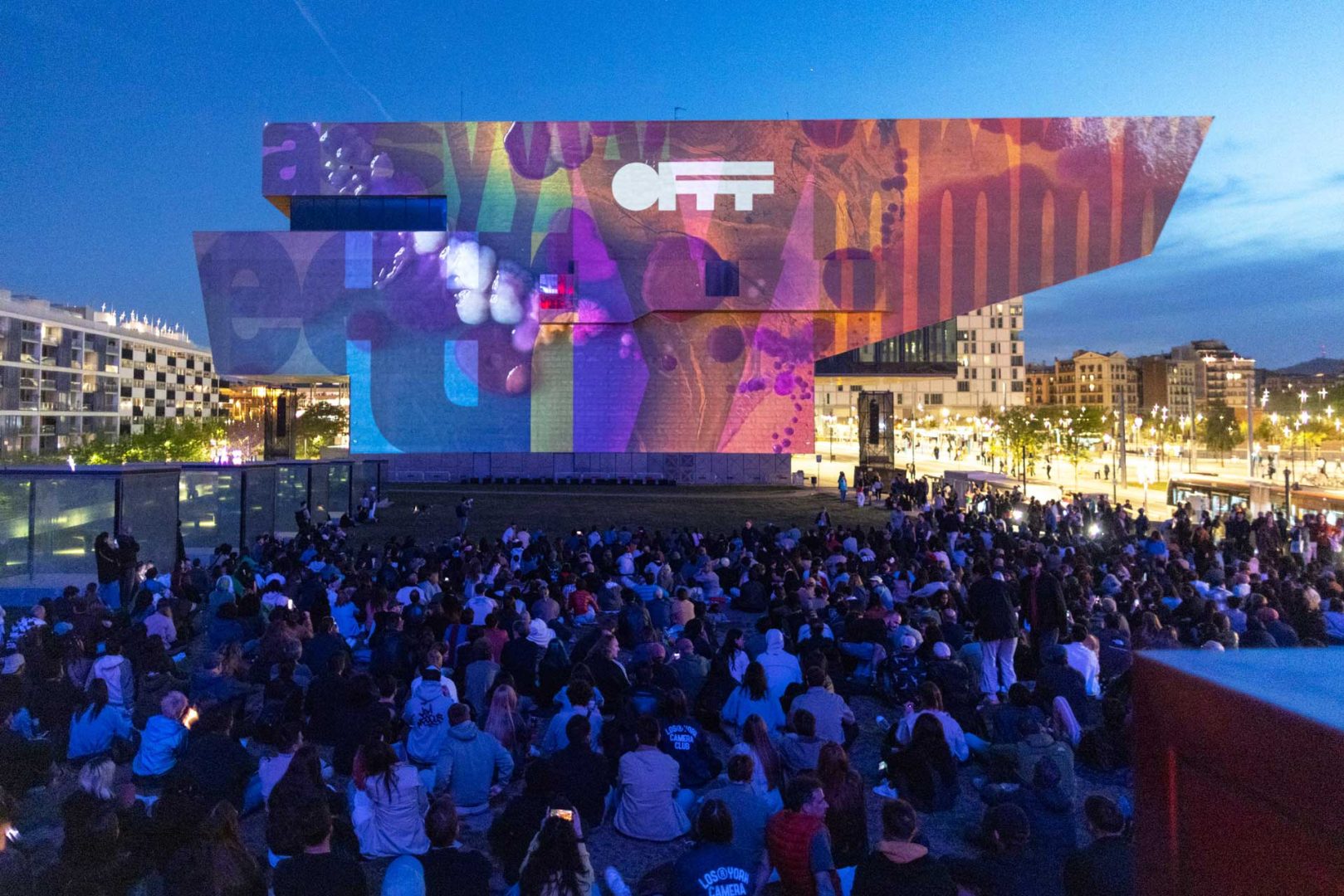 A large crowd sits on grass at dusk, watching the OFFF Barcelona 2026 Main Titles and colorful digital projections illuminate the modern, angular facade of a building in an urban setting. Frame from Stash Magazine article.