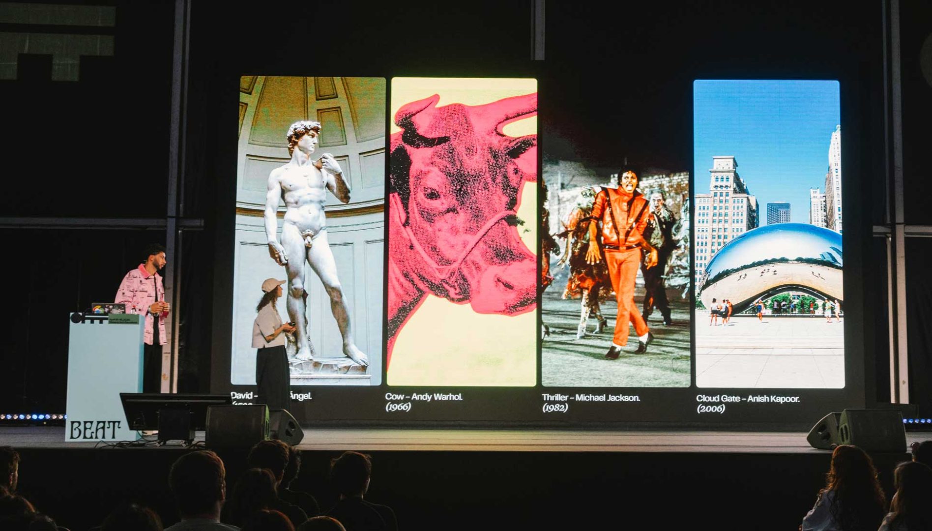 Valeria Moreiro, Creative Director at NotReal and Co-Founder, stands on stage beside a large screen displaying four famous artworks: Michelangelo’s David, Warhol’s Cow, Michael Jackson’s Thriller, and Anish Kapoor’s Cloud Gate. Frame from Stash Magazine article.